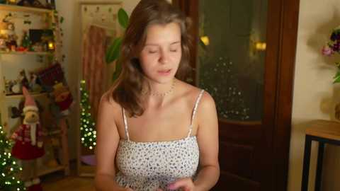 Media: Video of a young woman with shoulder-length brown hair, wearing a white polka-dot dress, standing indoors with Christmas decorations in the background.