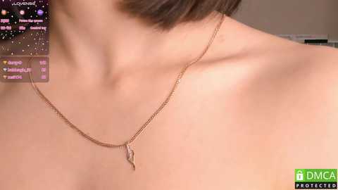 Media: Video of a close-up of a woman's bare shoulder, wearing a delicate gold necklace with a feather pendant, in a soft, dimly lit room.