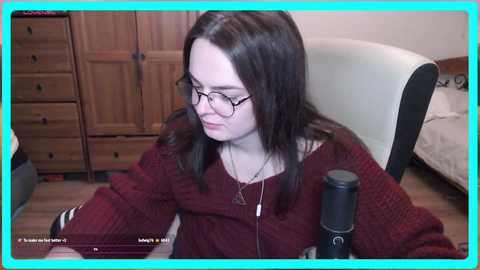 Media: Video of a young woman with fair skin, long dark hair, and glasses, wearing a burgundy sweater, sitting at a desk, surrounded by wooden furniture and a white chair.