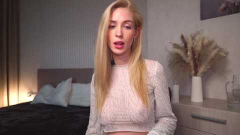 Media: Video of a blonde, slim woman in a white, ribbed crop top, seated on a bed in a modern bedroom. Background features a white wall, a potted plant, and a nightstand.