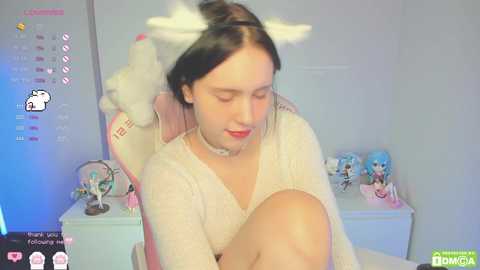 Media: A video of a young woman with fair skin, black hair, and a white cat headband, wearing a sheer white top, sitting in a room with a pink chair, blue wall, and plush toys.