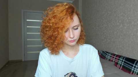 Media: Video of a young woman with curly red hair, wearing a white T-shirt, standing in a minimalist hallway with beige walls and a plaid blanket on a bench.