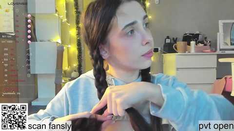 Media: A video shows a young woman with light skin and long dark hair, braided, in a casual light blue top, sitting at a desk. Background features a white desk, a calendar, and a pink handbag.