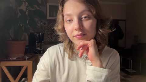 A video of a young girl with wavy, shoulder-length brown hair, light skin, and a neutral expression. She's wearing a white long-sleeve shirt and is seated in a dimly lit room with a potted plant, a wooden table, and a chair in the background.