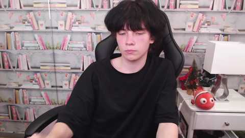 Media: Video of a pale, young man with dark hair, wearing a black shirt, seated in a black gaming chair, surrounded by colorful bookshelves and a red toy.