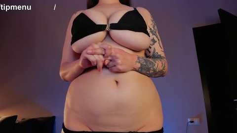 Media: Video of a plus-sized woman with large breasts, dark hair, and extensive tattoos, wearing a black bra, standing in a dimly lit room.