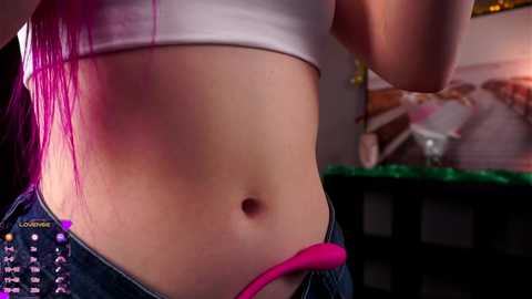 Video of a woman's midriff, showing a pink vibrator partially inserted into her unbuttoned jeans. She has a white crop top and long, straight, pink hair.