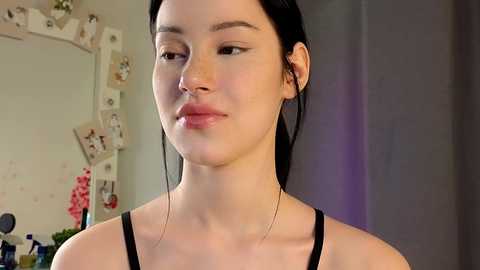 Media: Video of a young East Asian woman with pale skin, dark hair pulled back, wearing a black spaghetti strap top. Background shows a cluttered room with a framed photo, wall art, and plants.