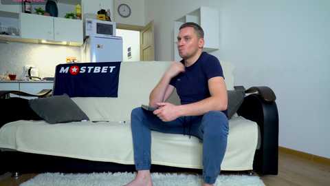 Media: Video of a young, fit, white man with short hair, wearing a navy t-shirt and blue jeans, sitting on a beige couch in a modern, minimalist kitchen with white walls and a blue \"MOSTBET\" tablecloth.