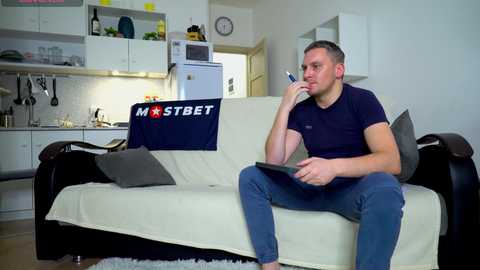 Media: Video of a clean, modern kitchen with white cabinets, a refrigerator, and a microwave. A man sits on a cream sofa, wearing a navy shirt, blue jeans, and holding a phone.