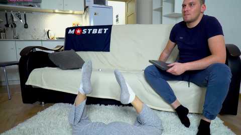 Media: Video of a man sitting on a cream couch, wearing a dark blue t-shirt and blue jeans, holding a tablet, while a person in pajamas lies on a white rug.