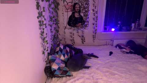 Media: Video of a dimly lit bedroom with a white bed covered in a patchwork quilt, a large wall tapestry, and a plush white rug.