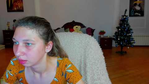 Media: Video of a young woman with light skin and blonde hair, wearing a colorful dress, looking pensive in a cozy, decorated bedroom with a Christmas tree and plush toys.