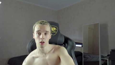 Media: A video of a young, shirtless, light-skinned man with short brown hair, sitting in a black gaming chair in a dimly lit room with beige walls and a mirror.