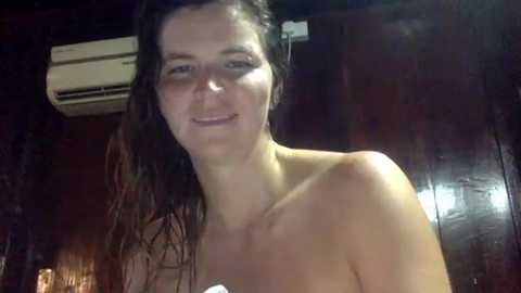 Media: Video of a topless woman with wet, dark hair, standing indoors with a dimly lit background featuring a wall-mounted air conditioner and wooden paneling.