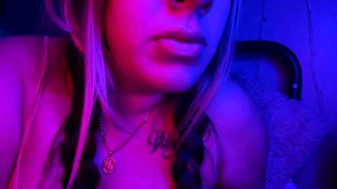 Media: Video of a young woman with fair skin and long blonde hair, wearing a necklace, under a purple and pink neon light.