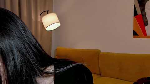 Media: A video shows a person with long, black hair, seated on a mustard-yellow sofa. A wall-mounted lamp with a cylindrical shade casts a warm light. The background features a beige curtain and a colorful abstract painting.