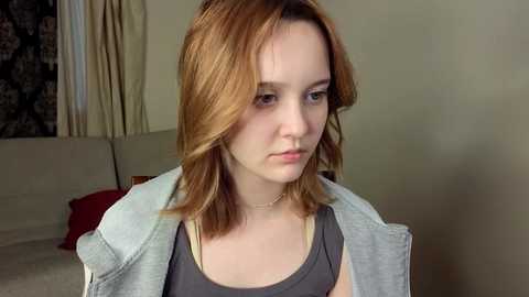 Media: Video of a young woman with shoulder-length, light brown hair, wearing a gray tank top and open light gray jacket, looking introspective in a beige room with a red pillow and abstract wall art.