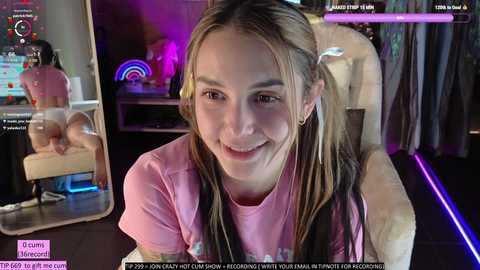 Media: Video of a smiling young woman with long brown hair, wearing a pink t-shirt, seated in a gaming chair in a dimly-lit room with neon lights.