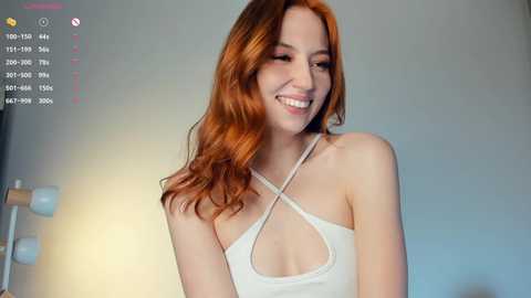 Media: Video of a smiling, red-haired woman with fair skin wearing a white, strappy top, standing in a softly lit room with a blurred background.