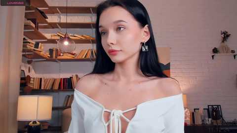 Video of a young woman with pale skin and long black hair, wearing a white off-shoulder top, standing indoors next to a bookshelf and modern light fixture.