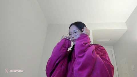 Media: A low-angle video of an Asian woman with glasses and a pink jacket, adjusting her glasses in a white-walled room.