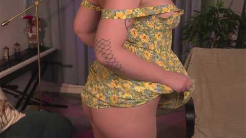 Media: Video of a plus-size woman in a yellow floral dress, exposing her side, revealing a geometric tattoo on her upper arm. Background includes a green chair, a lamp, and a plant.