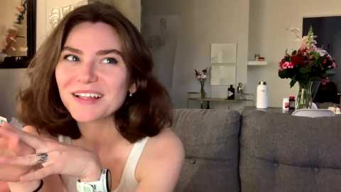 Media: Video of a smiling, fair-skinned woman with wavy brown hair, wearing a white tank top, gesturing excitedly in a modern living room with gray couch, floral arrangement, and framed artwork on the wall.