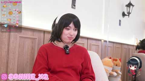 Media: Video of a young Asian woman with shoulder-length black hair, wearing a red sweater and a black choker, sitting indoors with a plush toy and a poster in the background.