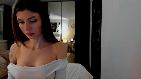 Media: Video of a fair-skinned woman with long brown hair, wearing a white off-shoulder top, looking down, in a dimly lit bedroom with mirrored closet doors and a bed with a textured quilt.