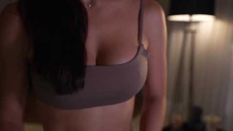 Media: Video of a woman with medium brown skin, wearing a gray sports bra, her dark hair cascading over her shoulder. Background features a dimly lit room with a lamp and blurred objects.