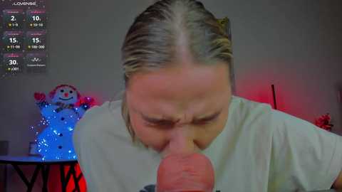 Media: A video shows a middle-aged woman with light skin, long blonde hair, and a white t-shirt performing oral sex on a man, who is partially visible. A festive snowman decoration and a digital display with stats are in the background.