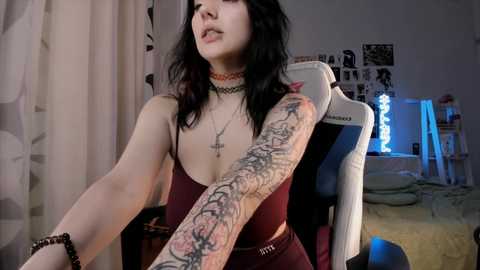 Media: Video of a pale-skinned woman with long black hair, wearing a maroon top and choker, displaying a detailed arm tattoo in a dimly lit bedroom with a gaming chair.