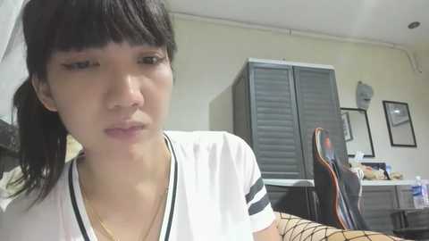 Media: A video of a young Asian woman with short black hair in pigtails, wearing a white T-shirt with black trim, looking contemplative in a cluttered, beige-walled room with gray cabinets and framed pictures.