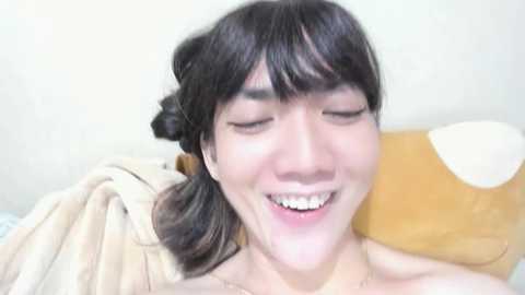 Media: Video of a smiling Asian woman with dark hair in a bun, wearing a beige top, lying on a beige bed with a stuffed animal.