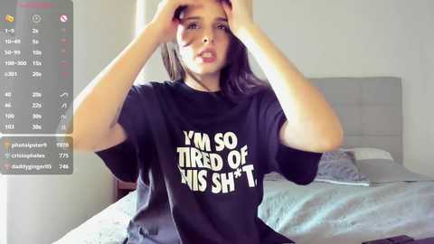 Media: Video of a young woman with light skin, long brown hair, and a medium build, wearing a black T-shirt with white text reading \"I'M SO TIRED OF HIS SHIT.\" She's sitting on a bed with grey pillows, in a brightly lit room.