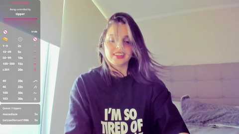 Media: Video of a young woman with shoulder-length, dark brown hair wearing a black hoodie with \"I'm so tired of\" text, smiling in a modern, well-lit bedroom.