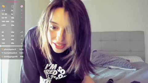 Media: Video of a young woman with long, straight brown hair, wearing a black T-shirt with white text, smiling in a bedroom with a grey upholstered headboard.