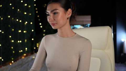 Media: Video of a young East Asian woman with medium skin tone, dark hair tied back, wearing a sheer, beige long-sleeve top, standing indoors beside a white chair, with a dark curtain and string lights in the background.