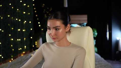 Media: Video of a young woman with light skin, dark hair in a bun, wearing a beige top, sitting on a white chair in a dimly lit room with fairy lights and greenery in the background.