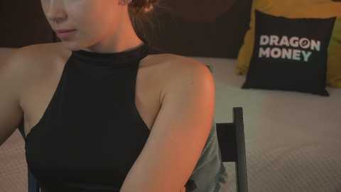 Media: Video of a woman with light skin, brown hair tied back, wearing a black halter top, sitting on a chair. Background features a black cushion with \"DRAGON MONEY\" text and a yellow pillow.