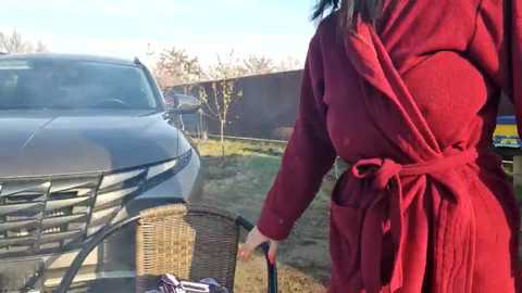 Media: Video of a woman in a red robe, standing next to a wicker chair, watering a plant in a garden. A gray car is parked in the background.