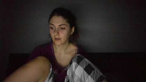 Media: A video of a young woman with dark hair pulled back, wearing a purple shirt and a black-and-white checkered blanket, sitting on a dark couch against a gray wall.