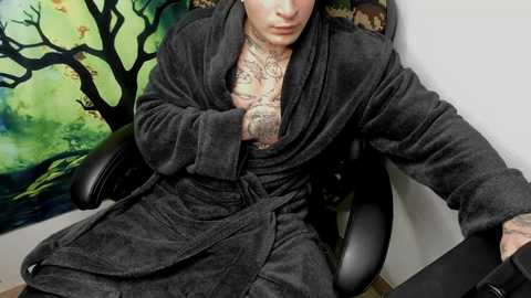 Media: A video of a tattooed, muscular man in a dark robe, sitting on a black office chair, with a colorful, abstract tree painting in the background.