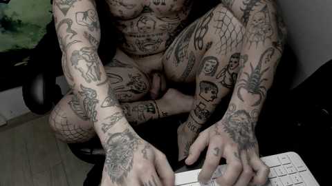 Media: Video of two naked tattooed men with diverse ink designs, including faces, spiders, and hearts, sitting on a black chair in a room with a green blanket and white tiles.