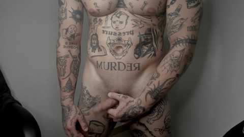 Media: Video of a nude, tattooed man with a muscular physique, covered in various designs including \"MURDER\" on his chest, holding a wooden cane, against a plain grey background.