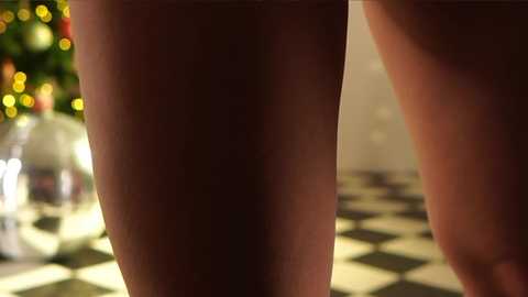 Media: A close-up video of a pair of legs in dim lighting, featuring a checkered floor and a blurred, festive background with a Christmas tree and ornaments. The focus is on the thighs, highlighting smooth, pale skin.