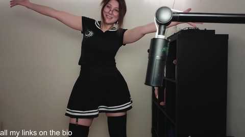 Media: Video of a cheerful woman in a black sailor dress and thigh-high stockings, adjusting a microphone stand against a beige wall, with text overlay \"all my links on the bio.\