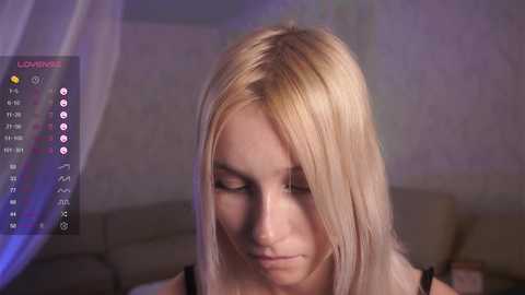 Media: Video of a blonde Caucasian woman with fair skin, wearing a black top, looking down, in a dimly lit room with a cream-colored couch and a white curtain.