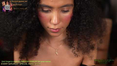 Media: A close-up video of a young woman with medium brown skin and large, voluminous curly hair, wearing a gold necklace. She has a contemplative expression, with subtle makeup, and is seen from the shoulders up.